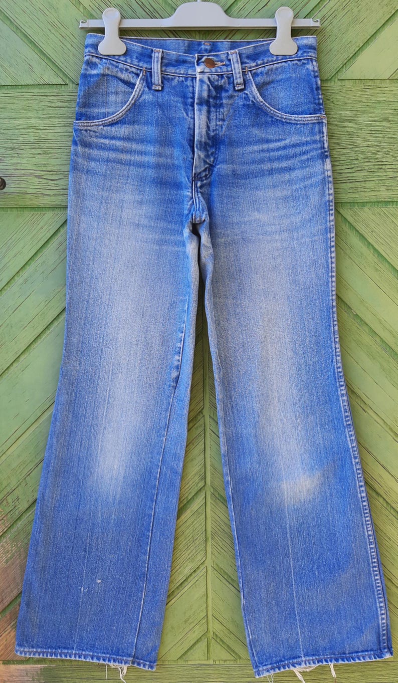 May include: Light blue denim jeans with a classic five-pocket design. The jeans have a high waist and a flared leg, with a faded wash and subtle wear marks. The jeans are hanging on a white hanger against a green background.