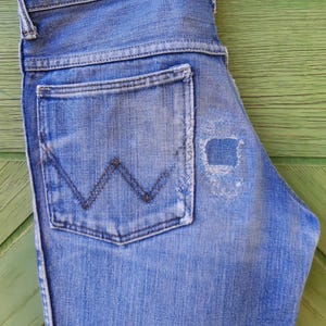 May include: A pair of blue denim jeans with a back pocket featuring a stitched W logo. The jeans have a distressed look with a small hole in the fabric.