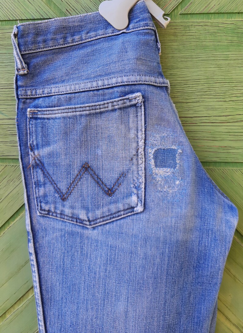 May include: A pair of blue denim jeans with a back pocket featuring a stitched W logo. The jeans have a distressed look with a small hole in the fabric.