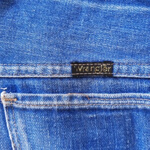 May include: Close-up of blue denim jeans with the brand name "Wrangler" on a black label. The jeans have a classic five-pocket design with visible stitching and a back pocket.