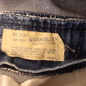 Pode incluir: Close-up da cintura de cal&ccedil;as de ganga Wrangler azuis. A etiqueta diz "30 X 31" e "925 DEN". As instru&ccedil;&otilde;es de lavagem tamb&eacute;m est&atilde;o impressas na etiqueta. As cal&ccedil;as de ganga s&atilde;o feitas nos EUA.