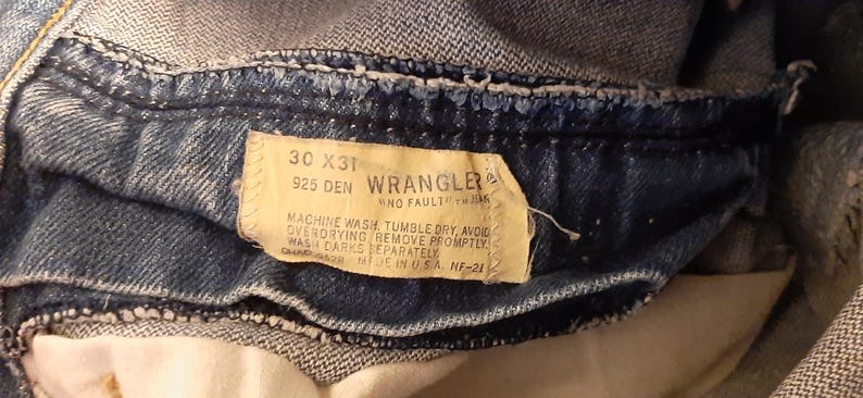 Pode incluir: Close-up da cintura de cal&ccedil;as de ganga Wrangler azuis. A etiqueta diz "30 X 31" e "925 DEN". As instru&ccedil;&otilde;es de lavagem tamb&eacute;m est&atilde;o impressas na etiqueta. As cal&ccedil;as de ganga s&atilde;o feitas nos EUA.