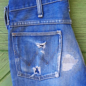 Pode incluir: Close-up de jeans de ganga azul com detalhes desgastados. Os jeans apresentam um bolso traseiro com um design cl&aacute;ssico e desgaste vis&iacute;vel. A ganga &eacute; de uma lavagem m&eacute;dia com &aacute;reas mais claras.