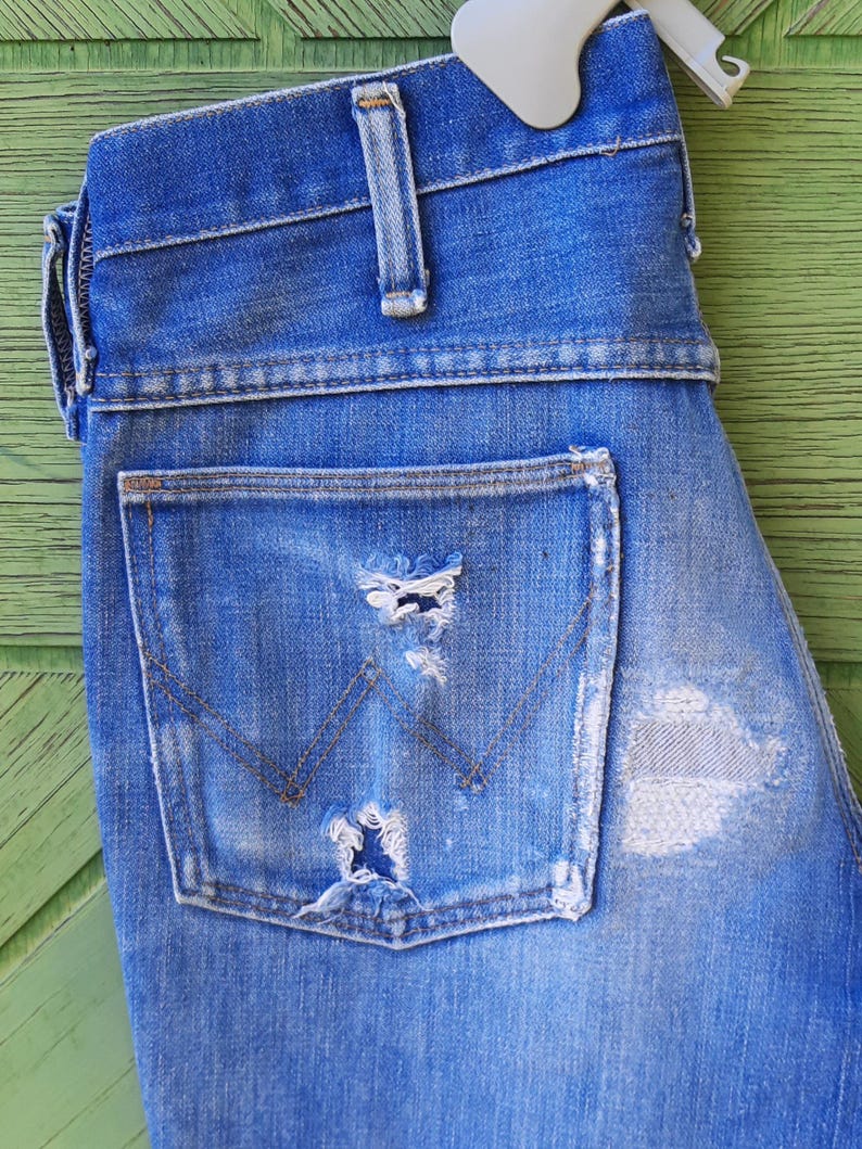 Pode incluir: Close-up de jeans de ganga azul com detalhes desgastados. Os jeans apresentam um bolso traseiro com um design cl&aacute;ssico e desgaste vis&iacute;vel. A ganga &eacute; de uma lavagem m&eacute;dia com &aacute;reas mais claras.