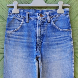 May include: Close-up of blue denim jeans hanging on a gray hanger. The jeans have a classic five-pocket design with a button and zipper closure. The denim has a faded wash, with lighter areas on the thighs and knees.