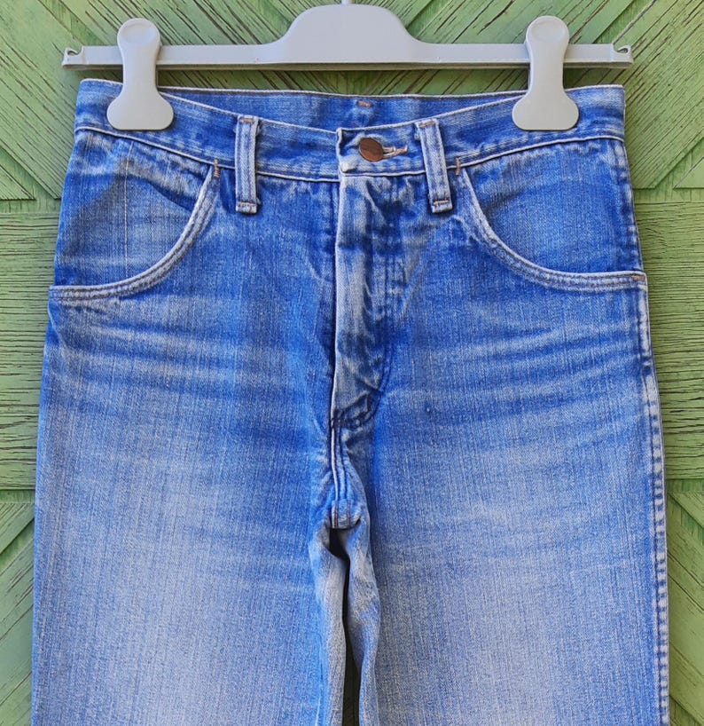 May include: Close-up of blue denim jeans hanging on a gray hanger. The jeans have a classic five-pocket design with a button and zipper closure. The denim has a faded wash, with lighter areas on the thighs and knees.