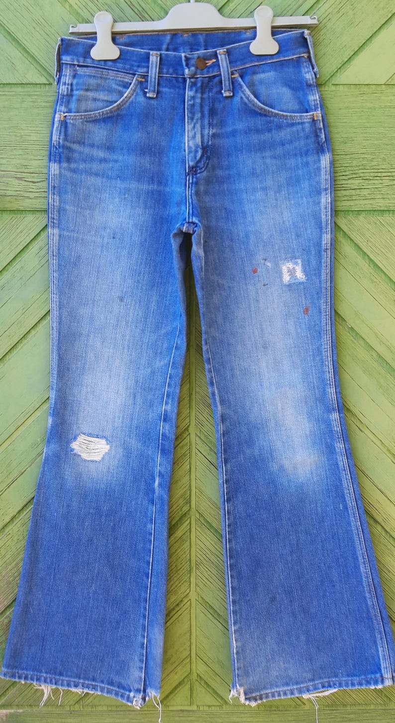 Pode incluir: Cal&ccedil;as de ganga azuis com perna larga, com joelho desgastado e &aacute;reas desbotadas. As cal&ccedil;as de ganga t&ecirc;m um design cl&aacute;ssico de cinco bolsos e est&atilde;o penduradas num cabide branco.