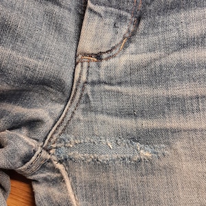 May include: Close-up of a pair of light blue denim jeans with a small tear in the fabric near the seam.