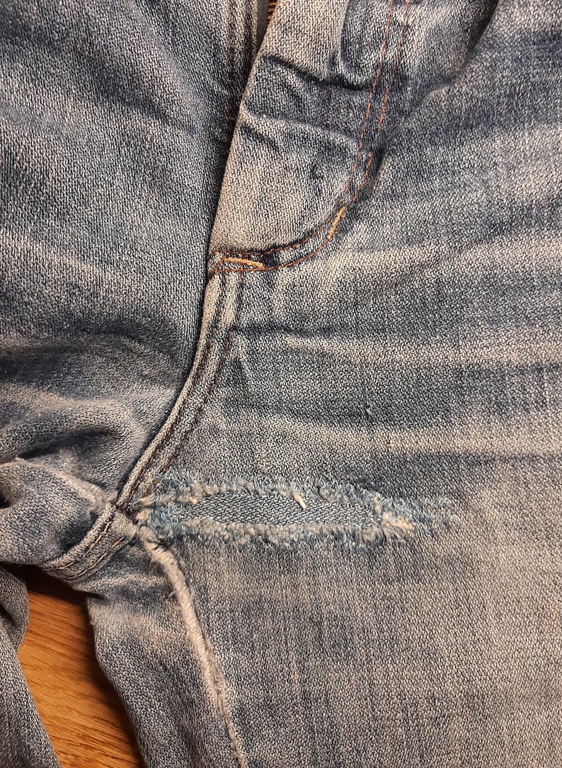 May include: Close-up of a pair of light blue denim jeans with a small tear in the fabric near the seam.