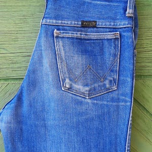 May include: Blue denim jeans with a Wrangler label on the back pocket, hanging from a white Prada hanger. The jeans have a classic five-pocket design and a faded wash. The background is a green wooden surface.