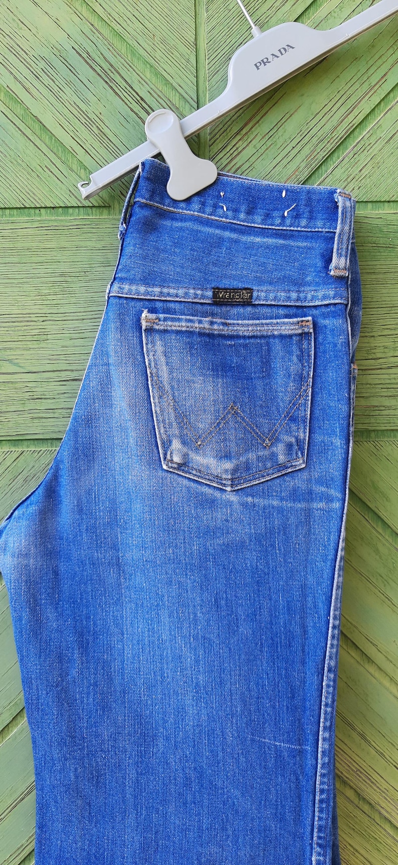 May include: Blue denim jeans with a Wrangler label on the back pocket, hanging from a white Prada hanger. The jeans have a classic five-pocket design and a faded wash. The background is a green wooden surface.
