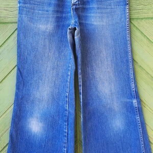 May include: Blue denim bell-bottom jeans hanging on a white hanger. The jeans have a button and zipper closure, front pockets, and a faded wash. The wide legs are a prominent feature of the vintage style.