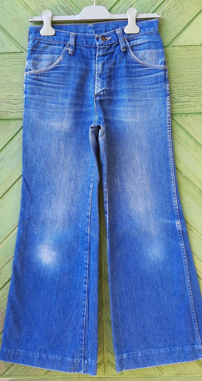 May include: Blue denim bell-bottom jeans hanging on a white hanger. The jeans have a button and zipper closure, front pockets, and a faded wash. The wide legs are a prominent feature of the vintage style.