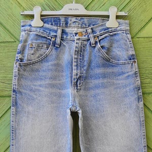 May include: Light blue denim jeans with a classic five-pocket design. The jeans have a faded wash and a straight leg cut. The jeans are hanging on a white hanger with the brand name "PRADA" printed on it.