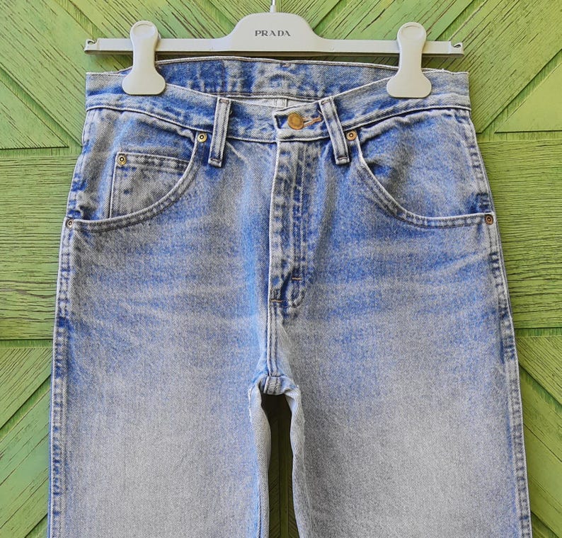 May include: Light blue denim jeans with a classic five-pocket design. The jeans have a faded wash and a straight leg cut. The jeans are hanging on a white hanger with the brand name "PRADA" printed on it.