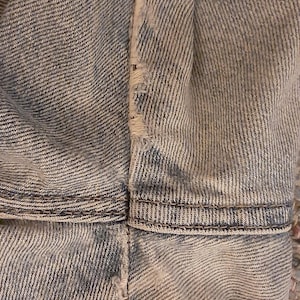 May include: Close-up of light blue denim fabric, showing the texture and stitching details. The image highlights the seams and the distressed edges of the denim, suggesting a worn or vintage style. The fabric appears to be from a pair of jeans or a denim jacket.