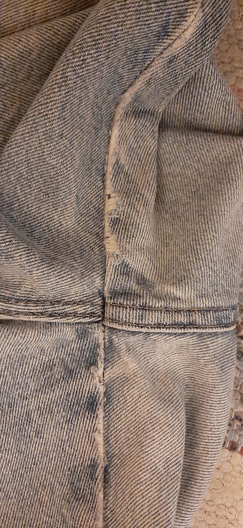 May include: Close-up of light blue denim fabric, showing the texture and stitching details. The image highlights the seams and the distressed edges of the denim, suggesting a worn or vintage style. The fabric appears to be from a pair of jeans or a denim jacket.