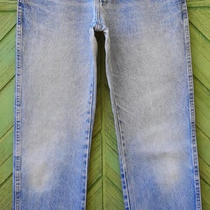 May include: Light blue denim jeans with a faded wash, hanging on a hanger. The jeans have a classic five-pocket design, a button and zipper closure, and a straight leg. The hanger has the brand name "PRADA" printed on it.