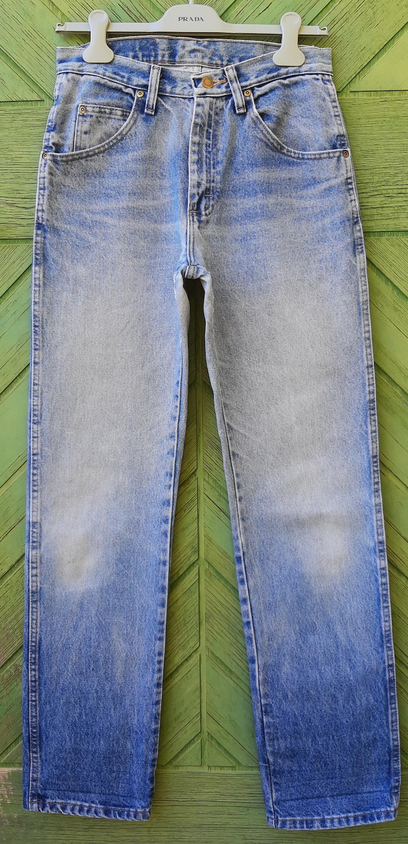 May include: Light blue denim jeans with a faded wash, hanging on a hanger. The jeans have a classic five-pocket design, a button and zipper closure, and a straight leg. The hanger has the brand name "PRADA" printed on it.