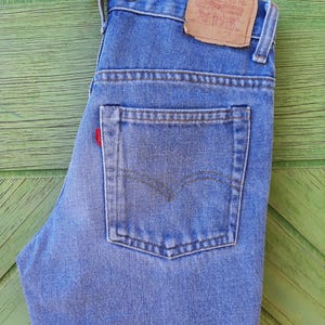 Può includere: Jeans blu in denim con un classico design a cinque tasche. I jeans presentano una toppa in pelle marrone sul retro con il nome e il logo del marchio. Un'etichetta rossa è visibile sulla tasca posteriore. I jeans sono appesi a una gruccia bianca.