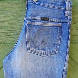 May include: Light blue denim jeans with a classic five-pocket design. The jeans feature a faded wash and a Wrangler brand label on the back pocket. The jeans are hanging on a white hanger against a green wooden background.