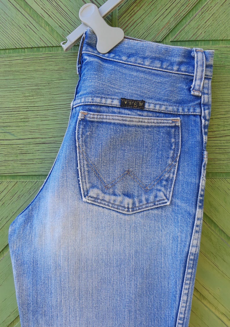 May include: Light blue denim jeans with a classic five-pocket design. The jeans feature a faded wash and a Wrangler brand label on the back pocket. The jeans are hanging on a white hanger against a green wooden background.