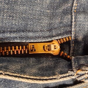 Pode incluir: Close-up de um par de jeans de ganga azul parcialmente deszipados. O fecho &eacute; de cor lat&atilde;o manchado e o bot&atilde;o &eacute; castanho escuro. O tecido de ganga mostra desgaste.