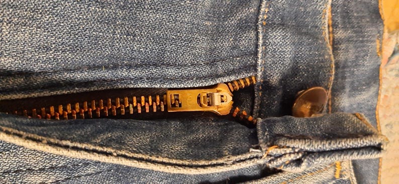 Pode incluir: Close-up de um par de jeans de ganga azul parcialmente deszipados. O fecho &eacute; de cor lat&atilde;o manchado e o bot&atilde;o &eacute; castanho escuro. O tecido de ganga mostra desgaste.