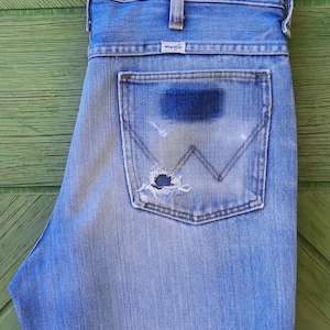 Może przedstawiać: Jasnoniebieskie dżinsy z przetartą kieszenią. Dżinsy mają klasyczny pięciokieszeniowy design, metkę Wrangler i zużytą naszywkę na tylnej kieszeni. Dżinsy wiszą na białym wieszaku na zielonym tle.