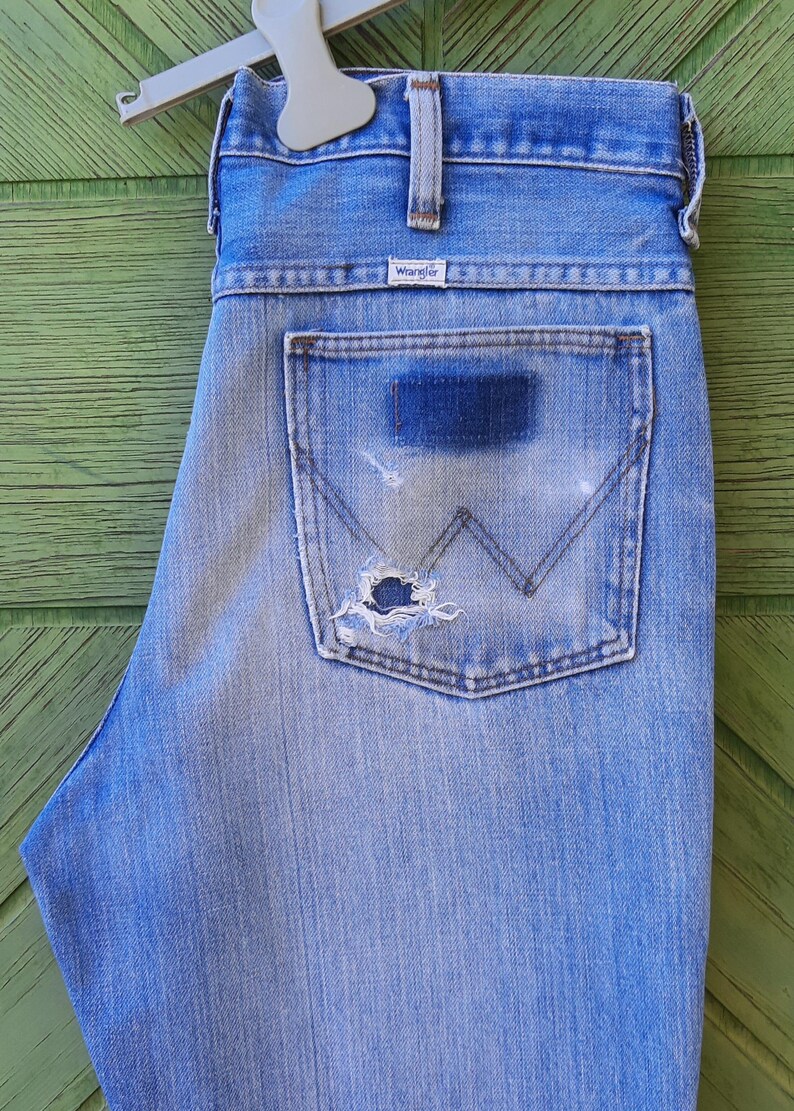 Może przedstawiać: Jasnoniebieskie dżinsy z przetartą kieszenią. Dżinsy mają klasyczny pięciokieszeniowy design, metkę Wrangler i zużytą naszywkę na tylnej kieszeni. Dżinsy wiszą na białym wieszaku na zielonym tle.