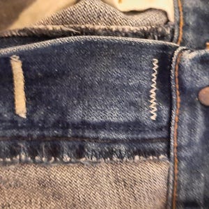 May include: Close-up of denim jeans showing the waistband, button, and stitching details. The blue denim fabric has a worn appearance, with visible seams and a metal button. The stitching is white.