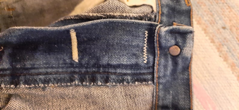 May include: Close-up of denim jeans showing the waistband, button, and stitching details. The blue denim fabric has a worn appearance, with visible seams and a metal button. The stitching is white.