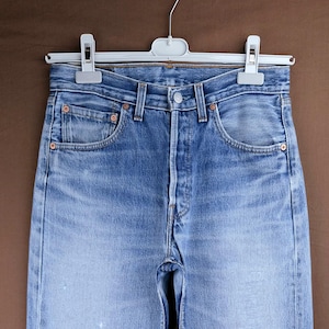 May include: Light blue denim jeans featuring a high-waisted cut, a button fly closure, and copper-toned metal rivets at the pocket corners. The fabric displays a faded wash with subtle whiskering details at the hips and a classic five-pocket design.