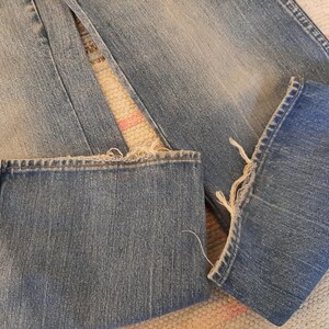 May include: Close-up of light blue denim jeans with frayed hems. The jeans are folded, showing the texture and stitching details. The fabric has a worn appearance, suggesting a vintage or distressed style.