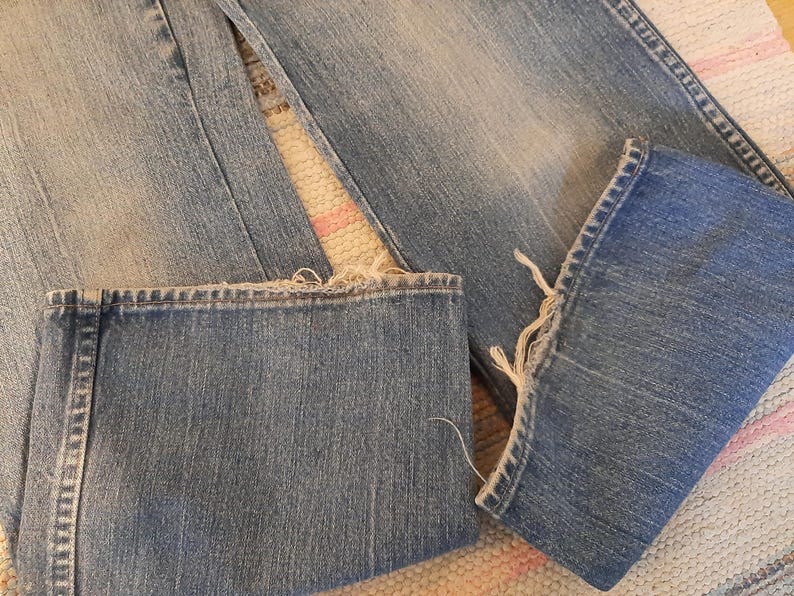 May include: Close-up of light blue denim jeans with frayed hems. The jeans are folded, showing the texture and stitching details. The fabric has a worn appearance, suggesting a vintage or distressed style.