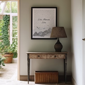 May include: A black framed print with the text "Live.  Manon.  Live."  The print is hanging on a light green wall above a wooden console table with a lamp and a wicker basket.