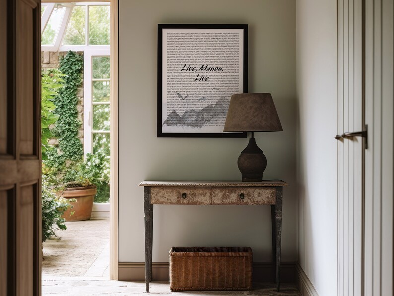 May include: A black framed print with the text "Live.  Manon.  Live."  The print is hanging on a light green wall above a wooden console table with a lamp and a wicker basket.