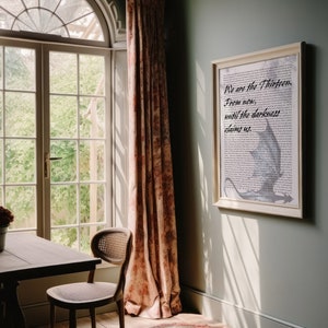 May include: A framed print with a white background and black text that reads "We are the Thirteen. From now, until the darkness claims us." The text is in a cursive font. A gray dragon is flying in the background. The print is hanging on a light green wall.