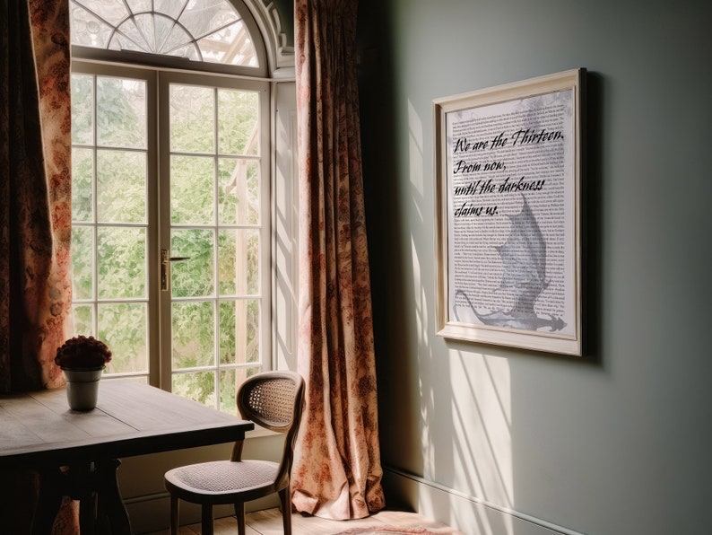 May include: A framed print with a white background and black text that reads "We are the Thirteen. From now, until the darkness claims us." The text is in a cursive font. A gray dragon is flying in the background. The print is hanging on a light green wall.