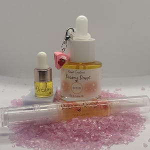 May include: Three bottles of Dreamy Drops cuticle and nail oil. The bottles are clear and contain a yellow liquid. The bottles have a white label with the text "Dreamy Drops" and "Cuticle & Nail Oil". The smallest bottle has a pink and white cake charm attached to the lid.