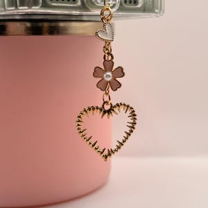 May include: A gold-colored phone charm with a heart, flower, and small heart design. The charm is attached to a clear phone case. The background is a light pink color.