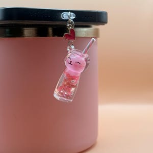 May include: A pink cat-shaped phone charm with a clear, liquid-filled body and a striped straw. The charm is attached to a phone and features a red heart charm. The charm is a cute accessory.