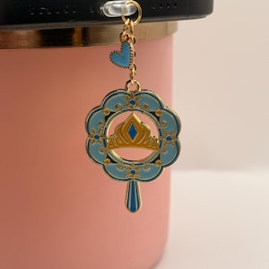 May include: A gold-colored phone charm with a blue and gold crown design. The charm is attached to a phone and features a small blue heart and a decorative floral-shaped pendant. The charm is hanging in front of a pink tumbler.