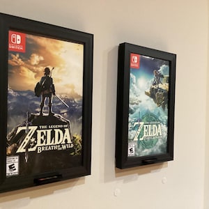 以下が含まれることがあります： Nintendo Switchのゲームカバーが2つ額装されています。黒い長方形のフレームには、「ゼルダの伝説 ブレス オブ ザ ワイルド」と「ゼルダの伝説 ティアーズ オブ ザ キングダム」のカバーアートが飾られています。カバーには、風景の中にいるキャラクターが描かれています。