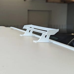 Könnte beinhalten: Ein kleiner, weißer, 3D-gedruckter Heckspoiler für Autos, der zur Montage an einem Fahrzeug bestimmt ist. Der Spoiler hat eine flügelartige Form mit zwei Stützpfeilern. Das Objekt befindet sich vor einem neutralen Hintergrund.