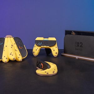 Op de afbeelding: Een verzameling Nintendo Switch-accessoires met een bananathema. Inclusief een dock, controllers en een Joy-Con-grip, allemaal in geel met bruine vlekken. Het dock is zwart met het Nintendo Switch-logo.