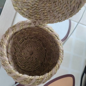 May include: A woven basket with a lid, crafted from natural fibers in shades of beige and brown. The basket's round shape and textured surface create a rustic aesthetic. The lid is partially open, revealing the interior of the basket.