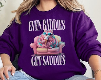 May include: Purple sweatshirt with a pink cat wearing sunglasses and eating ice cream on a pink couch. The text "Even Baddies Get Saddies" is printed on the sweatshirt.