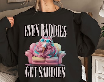 May include: Black sweatshirt with a pink and blue cat wearing sunglasses and eating ice cream. The text "Even Baddies Get Saddies" is printed on the shirt.