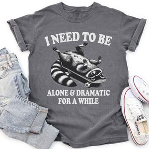 May include: A dark gray t-shirt with the text "I need to be alone & dramatic for a while" and a cartoon raccoon skateboarding.